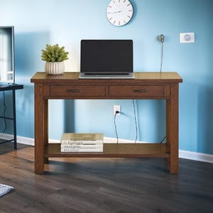 Puede incluir: Una mesa consola rectangular de madera con acabado marrón medio, que cuenta con dos cajones con tiradores de metal oscuro y un estante inferior. Un portátil se sitúa sobre la superficie y tres libros titulados Kinfolk, Vogue Living y Picasso.