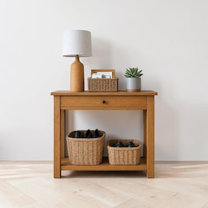 Puede incluir: Una mesa consola de madera con un cajón y un estante inferior. Encima hay una lámpara color canela con pantalla blanca, una cesta tejida pequeña y una suculenta. Dos cestas tejidas más grandes con zapatos negros descansan en el estante inferior.