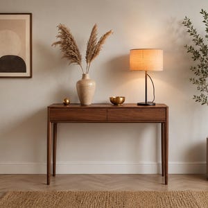 May include: A rectangular dark wood console table with two drawers and four slender legs. On the table sits a beige ceramic vase with dried pampas grass, a small gold sphere, a gold bowl, and a table lamp with a cylindrical beige fabric shade.