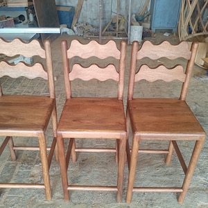 Op de afbeelding: Drie houten stoelen met een golvend ontwerp op de rugleuningen. De stoelen hebben vierkante zittingen en vier rechte poten. Het hout heeft een warme, roodbruine tint. De stoelen zijn naast elkaar geplaatst.