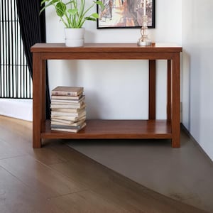 Puede incluir: Una mesa consola rectangular de madera con un acabado marrón cálido, que presenta una superficie superior plana y un estante inferior. Una pila de diez libros descansa sobre el estante inferior, mientras que una planta verde en maceta está arriba.