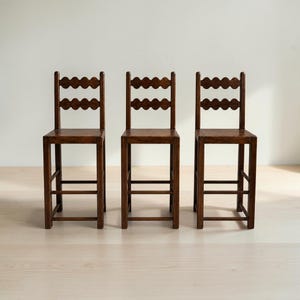 Op de afbeelding: Drie hoge houten barkrukken met een donkerbruine afwerking. Elke kruk heeft een vierkante zitting, vier rechte poten met horizontale steunbalken en een rugleuning bestaande uit twee horizontale latten met een geschulpte rand.