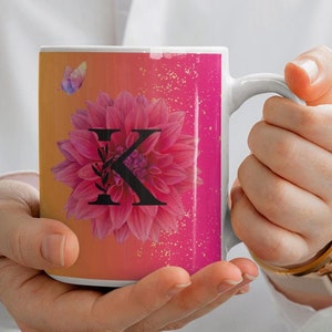 May include: White ceramic mug with a pink and orange ombre design. The mug features a pink flower with a black letter "X" in the center. A small butterfly is in the upper left corner of the design.