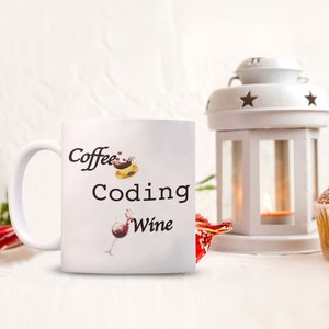 Pode incluir: Caneca de cer&acirc;mica branca com as palavras "Coffee Coding Wine" em escrita preta. A caneca apresenta ilustra&ccedil;&otilde;es de uma x&iacute;cara de caf&eacute;, um s&iacute;mbolo de codifica&ccedil;&atilde;o de computador e uma ta&ccedil;a de vinho. Uma lanterna branca e um item decorativo vermelho est&atilde;o no fundo.