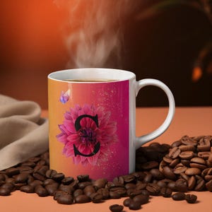 May include: White ceramic mug with a pink and orange ombre design. The mug features a pink flower with a black letter "S" in the center. The mug is surrounded by coffee beans.