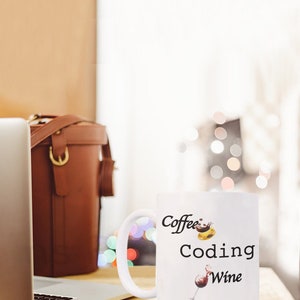 Pode incluir: Caneca de cer&acirc;mica branca com as palavras "Coffee Coding Wine" em escrita preta. A caneca apresenta ilustra&ccedil;&otilde;es de caf&eacute;, um computador e uma ta&ccedil;a de vinho. Um laptop e uma bolsa de couro marrom est&atilde;o no fundo.