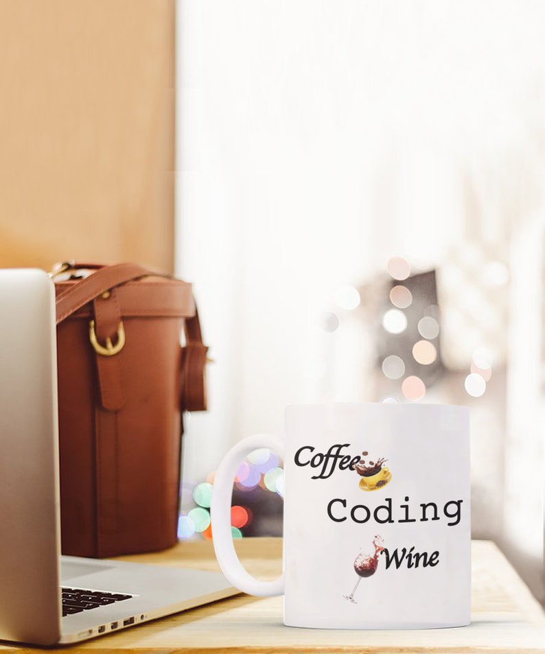 Pode incluir: Caneca de cer&acirc;mica branca com as palavras "Coffee Coding Wine" em escrita preta. A caneca apresenta ilustra&ccedil;&otilde;es de caf&eacute;, um computador e uma ta&ccedil;a de vinho. Um laptop e uma bolsa de couro marrom est&atilde;o no fundo.