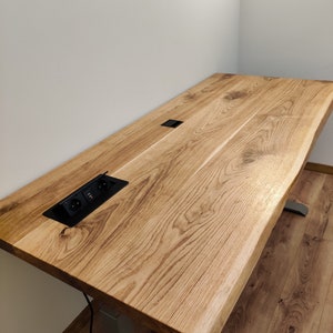 Könnte beinhalten: Ein Holzschreibtisch mit einer Live-Edge-Oberfläche und einer integrierten Steckdose. Der Schreibtisch ist aus hellbraunem Holz mit einer natürlichen Oberfläche. Der Schreibtisch steht auf einem verstellbaren Untergestell aus Silber.