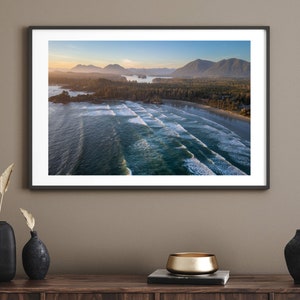 May include: An aerial photograph of a beach with waves crashing on the shore. The beach is surrounded by lush green trees and mountains in the distance. The sky is a light blue with a few clouds.