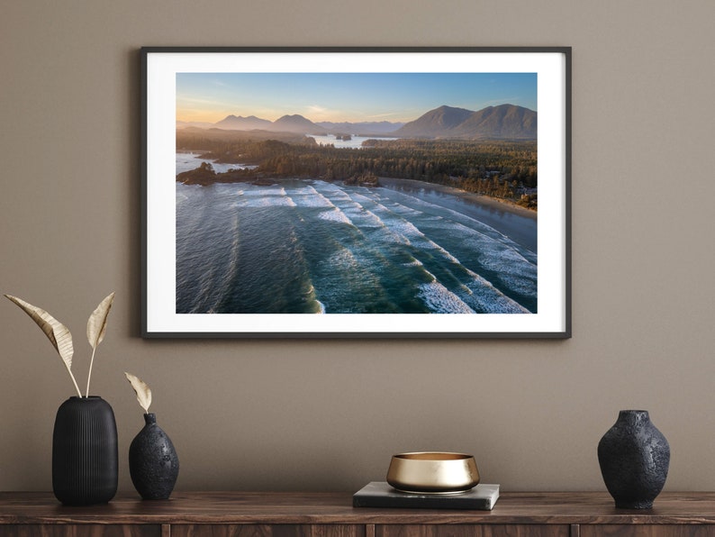 May include: An aerial photograph of a beach with waves crashing on the shore. The beach is surrounded by lush green trees and mountains in the distance. The sky is a light blue with a few clouds.