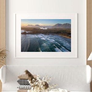 May include: An aerial photograph of a beach with waves crashing on the shore. The beach is surrounded by lush green trees and mountains in the distance. The sky is a light blue with a few clouds.