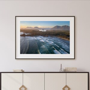 May include: An aerial photograph of a beach with waves crashing on the shore. The beach is lined with trees and a small town in the distance. The sky is a light blue with a few clouds.