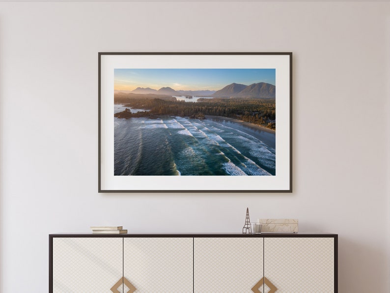 May include: An aerial photograph of a beach with waves crashing on the shore. The beach is lined with trees and a small town in the distance. The sky is a light blue with a few clouds.