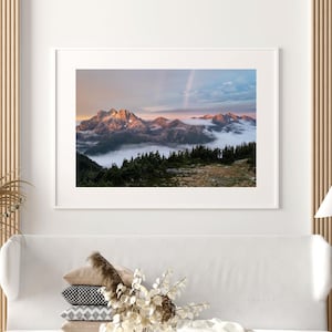 Könnte beinhalten: Ein Foto einer Bergkette mit einer Wolkendecke, die die unteren Hänge bedeckt. Die Berge werden vom untergehenden Sonnenlicht beleuchtet, das einen warmen Schein auf die Gipfel wirft. Der Himmel ist eine Mischung aus Blau und Rosa, mit einem schwach sichtbaren Regenbogen in der Ferne.