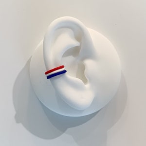 May include: A red, white, and blue ear cuff is displayed on a white ear model. The cuff is a simple band design, with the colors arranged in stripes. The ear model is smooth and white, providing a clean backdrop for the jewelry.
