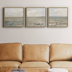May include: Three framed seascape paintings with gold frames. The paintings depict a muted color palette of blues, greens, and grays, with a focus on the ocean and sky. The artwork is displayed above a tan leather sofa.