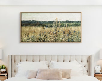 May include: A framed print of a field landscape with tall grasses and wildflowers in gold, green, and blue hues. The artwork, with a neutral palette, is displayed above a bed with a beige headboard. The frame is a light wood.
