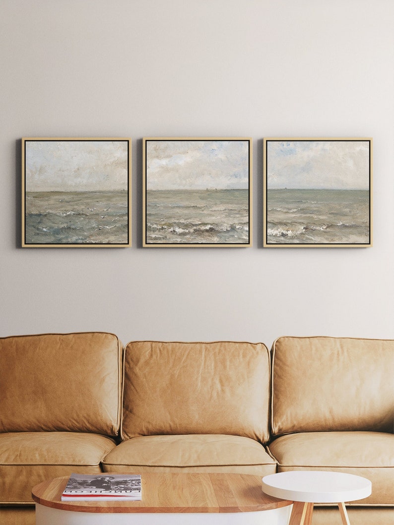 May include: Three framed seascape paintings with gold frames. The paintings depict a muted color palette of blues, greens, and grays, with a focus on the ocean and sky. The artwork is displayed above a tan leather sofa.