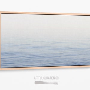 May include: A rectangular canvas print featuring a serene seascape. The artwork depicts a gradient of colors, transitioning from a light, misty sky to a calm, blue ocean. The canvas is framed in a natural wood border, adding a touch of warmth to the cool tones of the artwork.