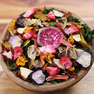 May include: A wooden bowl filled with a colorful assortment of dehydrated vegetable and fruit pieces, including bright pink radish slices, dark green kale, yellow cauliflower florets, red strawberry bits, and various other dried produce.