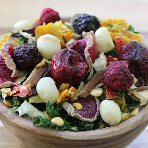 May include: A wooden bowl filled with a mixture of dried food items, including dark red cherries, white macadamia nuts, thin slices of purple and red dried vegetables, and green leafy pieces, creating a textured and colorful assortment of snacks.