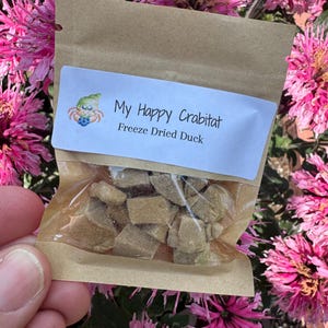 May include: A small brown paper pouch with a clear plastic window containing freeze-dried duck pieces. The white label features a hermit crab illustration and the text My Happy Crabat Freeze Dried Duck in black font on a white background.