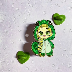 以下が含まれることがあります： 金色のエナメルピン。緑のカエルのコスチュームを着た漫画のキャラクターが描かれています。キャラクターは長い金髪で、笑顔です。2つの緑色のハート型のピンバックも表示されています。