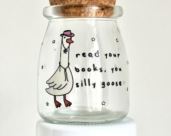 May include: A clear glass jar with a cork lid and a hand-painted illustration of a goose wearing a cowboy hat. The jar has the text "read your books, you silly goose" written on it.