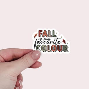 May include: A sticker with the text "FALL is my favourite COLOUR" in a mix of colours including red, green, and orange. The sticker has a white border and is held by a hand against a light pink background. The sticker has a fall theme.