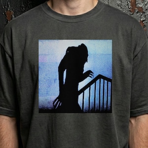 Puede incluir: Camiseta gris oscuro con un gráfico cuadrado que representa una silueta negra de una figura en una escalera. La figura tiene manos con forma de garra y una postura encorvada, sobre un fondo azul y blanco. La camiseta es de cuello redondo.