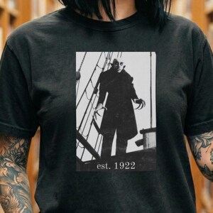 May include: Black t-shirt featuring a black and white image of a vampire from the 1922 film Nosferatu. The image shows the vampire standing on a ship with the text "est. 1922" below the image. The shirt is a short-sleeved crew neck.