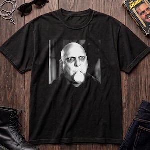 May include: A black short-sleeved t-shirt featuring a centered, square, black-and-white graphic print of a bald individual with wide, staring eyes and a round object held in their mouth. The shirt has a crew neckline and a standard, relaxed fit.