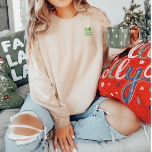 Op de afbeelding: Een vrouw draagt een lichtbeige sweatshirt met de tekst "cdi winter" in groene letters. Ze zit op een bank met een rood en wit kussen waarop "holly" in roze letters staat. Ze houdt een groene mok vast met een wit ontwerp.