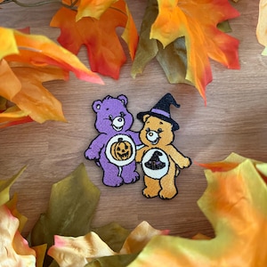 May include: Two embroidered patches featuring cartoon bears. One purple bear holds a pumpkin, and one orange bear wears a black witch hat. The orange bear also has a black witch hat icon on its white belly patch, while the purple bear has a pumpkin icon.