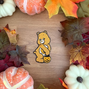 May include: An embroidered patch featuring a yellow bear character with a white belly patch, holding a small black jack-o'-lantern bucket. The bear has a black outline, stitched details on its face, and a small tail, presented as a flat textile item.