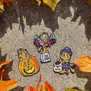 May include: Three embroidered patches featuring the cartoon character Garfield in Halloween costumes. One shows Garfield in a pumpkin, one as a vampire standing on a tombstone labeled TV DAD, and one as a pirate holding a bag labeled TRICK OR TREAT.