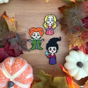 May include: Three embroidered patches featuring stylized cartoon characters. One character has bright orange hair and a green dress, one has long blonde hair and a pink dress, and one has black hair in a tall style and a purple dress.