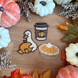 May include: Three embroidered patches featuring a white duck holding an orange pumpkin, a brown coffee cup with a pumpkin icon, and a slice of pumpkin pie with whipped cream, all with black outlines, arranged on a wooden surface with pumpkins.