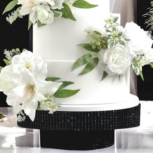 Pode incluir: Um bolo branco de três camadas adornado com arranjos florais brancos e folhagem verde. O bolo repousa sobre uma base preta com lantejoulas. O bolo é decorado com grandes flores brancas e flores brancas menores e vegetação.