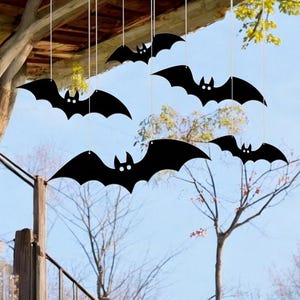 May include: Black bat decorations hanging from strings against a blue sky. The bats have white eyes and are suspended from a wooden structure and tree branches, creating a spooky Halloween scene.
