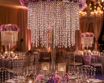May include: Elegant table setting with a centerpiece featuring a floral arrangement of pink and purple flowers atop a crystal chandelier. The table is set with silver chargers, glassware, and candles, creating a luxurious ambiance.