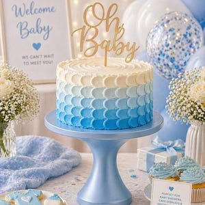 May include: A baby shower cake with an ombre blue design, topped with a gold "Oh Baby" cake topper. The cake sits on a blue cake stand, surrounded by balloons, cupcakes, and cookies. A sign reads "Welcome Baby".
