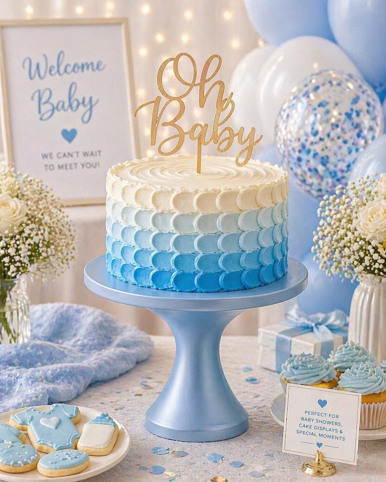 May include: A baby shower cake with an ombre blue design, topped with a gold "Oh Baby" cake topper. The cake sits on a blue cake stand, surrounded by balloons, cupcakes, and cookies. A sign reads "Welcome Baby".