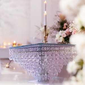 Könnte beinhalten: Ein quadratischer Glaskuchenständer mit einem kristallartigen Perlenbesatz. Der Ständer ist klar und hat einen Perlenbesatz an den Rändern. Der Ständer steht auf einer weißen Oberfläche.