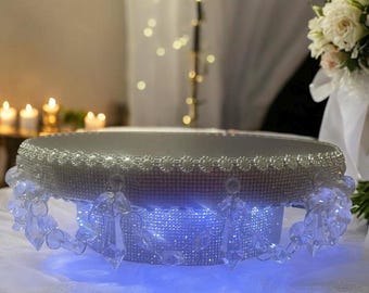 May include: An ornate cake stand with a mirrored silver top and a base adorned with shimmering rhinestones. It has hanging crystal embellishments and is lit with blue lights, creating an elegant and eye-catching presentation.