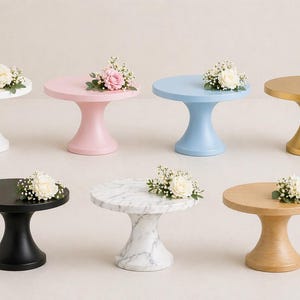 May include: A collection of small, decorative cake stands in various colors and materials. The stands include white, pink, blue, and gold painted finishes, as well as black, marble, and wood. Each stand is topped with a small floral arrangement.