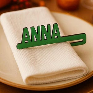 Può includere: Un segnaposto in legno verde con il nome "ANNA" ritagliato, appoggiato su un tovagliolo bianco. Il tovagliolo è su un piatto color crema con bordo dorato, che suggerisce un'apparecchiatura.