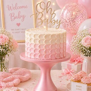 May include: A two-tiered cake with a pink and white ombre design, topped with a gold glitter "Oh Baby" cake topper. The cake sits on a pink cake stand, surrounded by pink balloons, flowers, and baby-themed cookies. A sign reads "Welcome Baby".