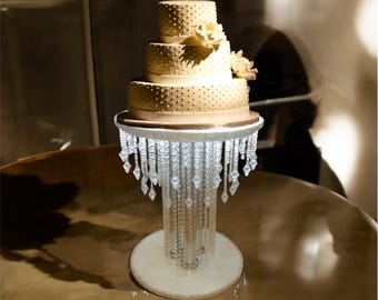 May include: A four-tiered wedding cake with white frosting and gold accents sits on a crystal cake stand with a chandelier-style design.