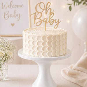 May include: A white frosted cake with a gold "Oh Baby" cake topper sits on a white cake stand. A framed sign that says "Welcome Baby" is in the background. The cake is decorated with a textured pattern.
